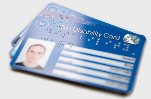 Drie European Disability Cards liggen op elkaar gestapeld. De bovenkant van de bovenste kaart is 'geblurred' zodat foto en gegevens niet afgelezen kunnen worden. De kaarten zijn donkerblauw, met links een pasfoto en rechts witte invulboxen. Op de bovenkant staat eveneens 'EU Disability Card' in Brailleletters.