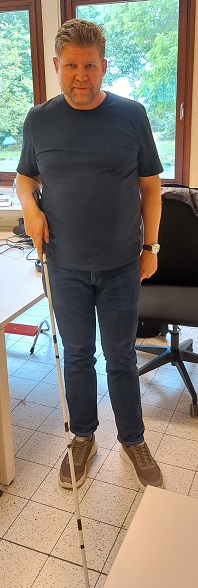 Een man met visuele Handicap demonstreert de Witte Stok; hij houdt de stok voor zich met de rechterhand. Hij draagt een blauwgrijze broek en T-shirt.